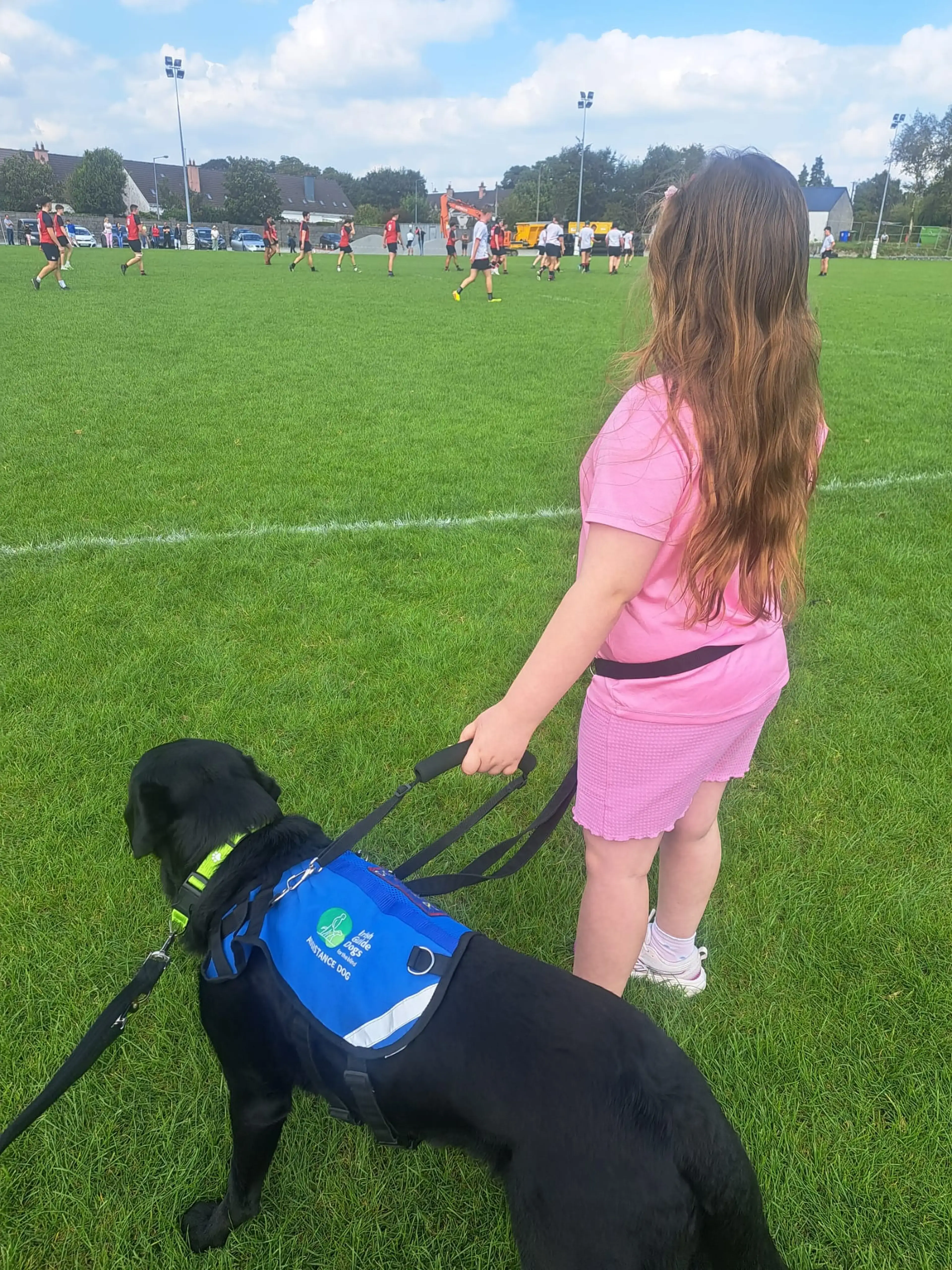 A girl in pink athletic wear stands on a soccer field with a black service dog wearing a blue vest, watching a youth soccer match in the background.