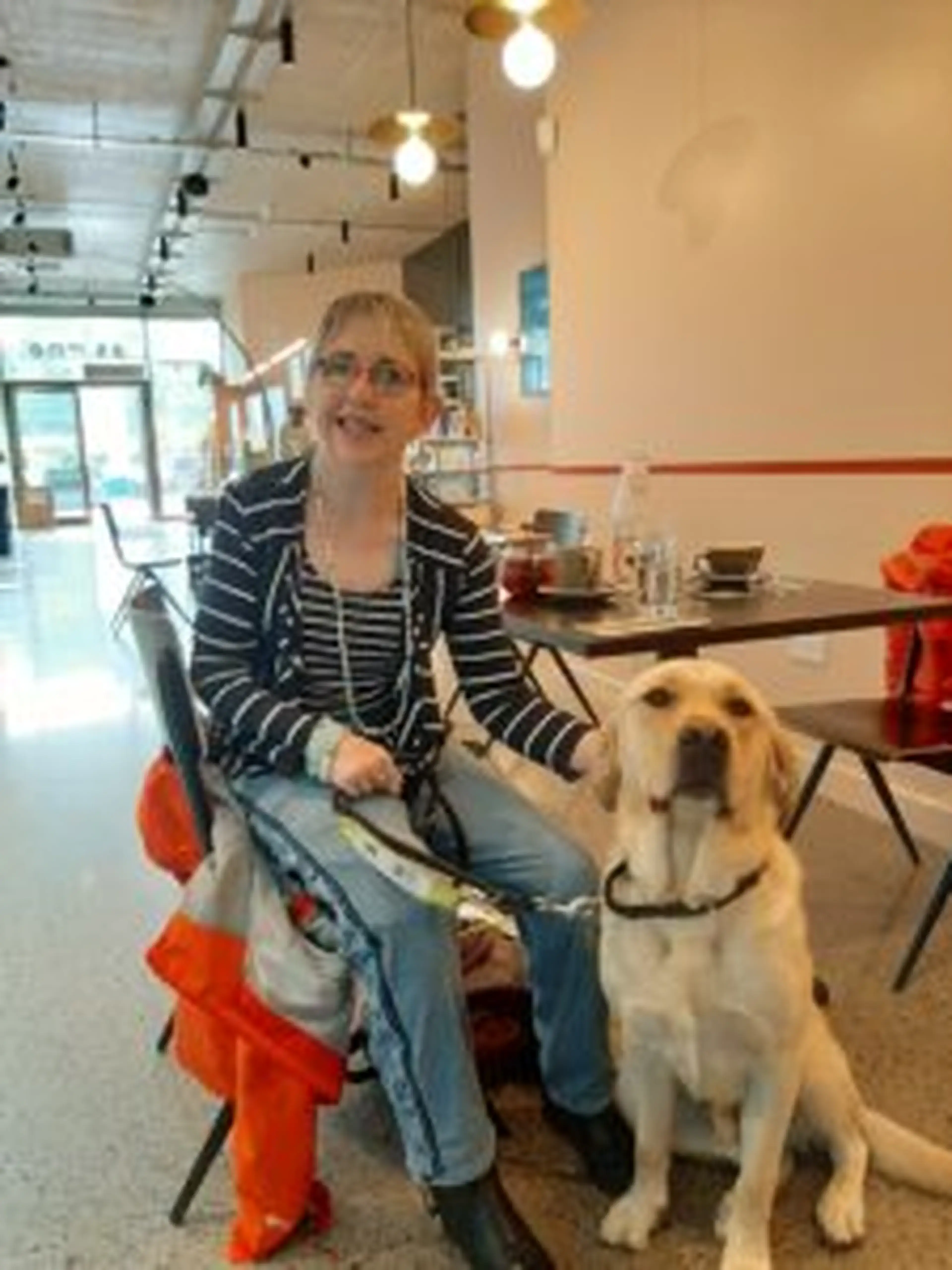 A woman wearing glasses and a striped jacket sits beside a yellow Labrador service dog in a modern indoor facility with track lighting and red safety equipment visible in the backg
