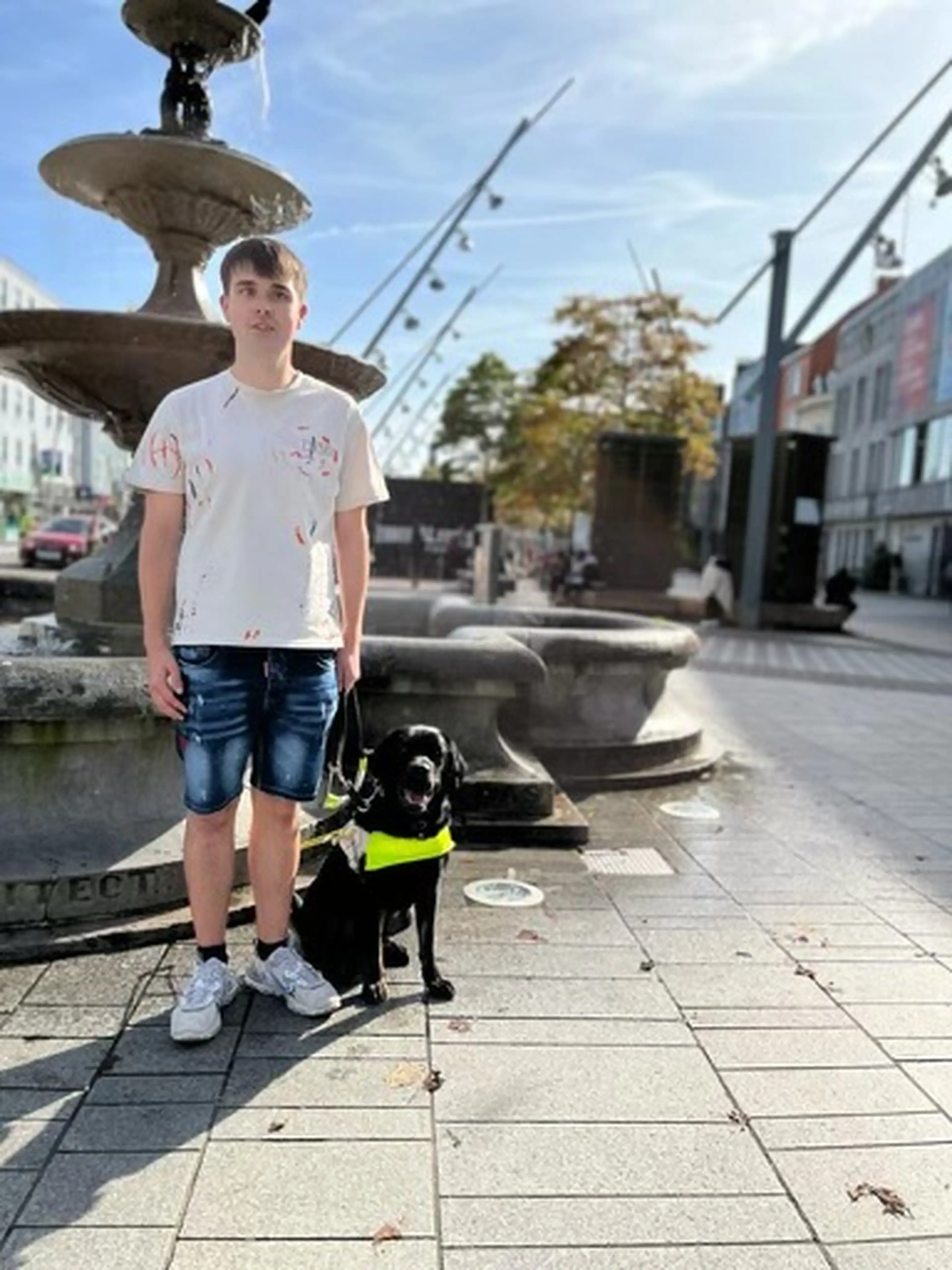 A young person stands with a service dog wearing a yellow harness in front of a large ornamental fountain in an urban plaza.