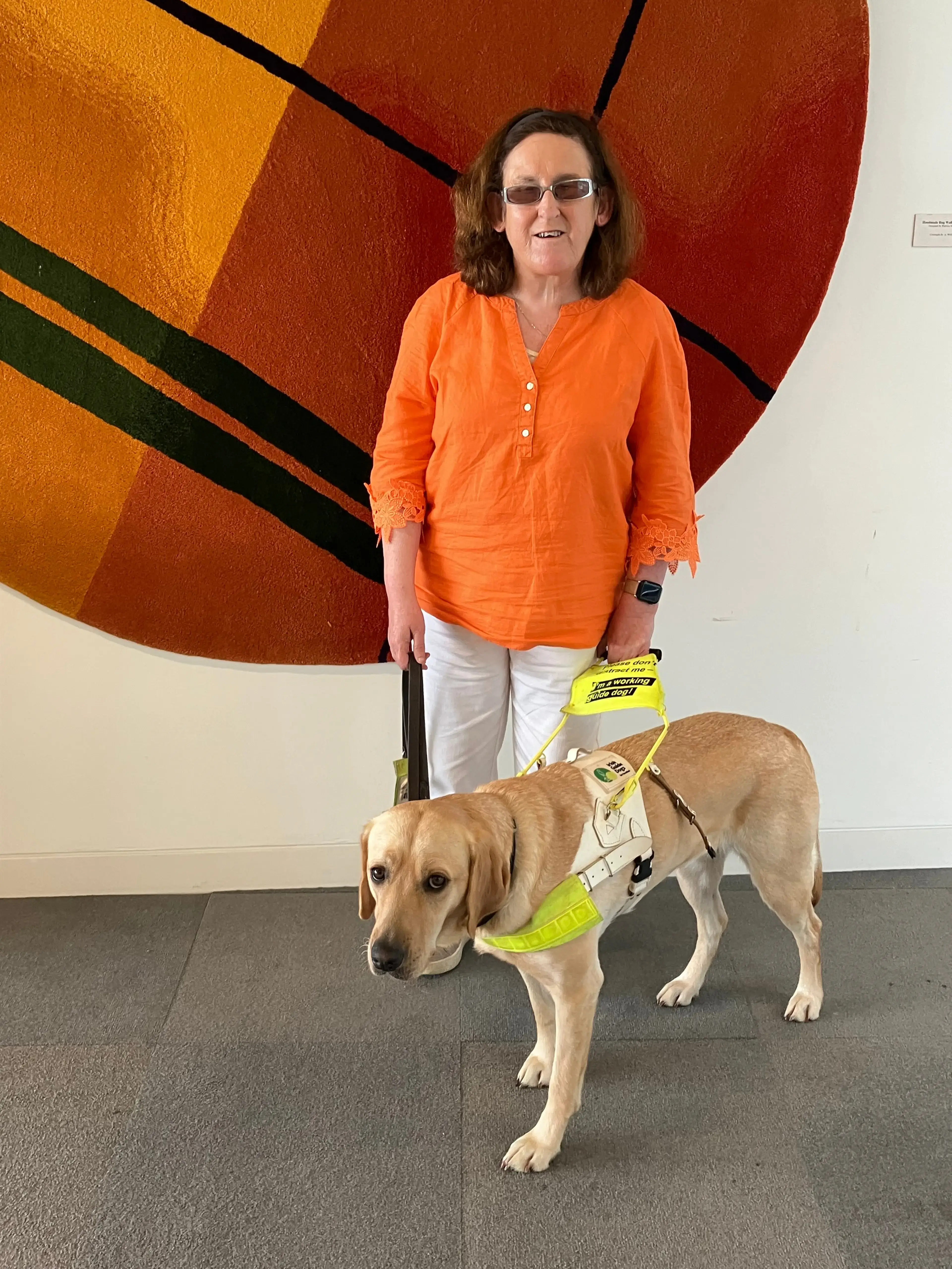 A woman wearing an orange shirt and sunglasses stands with a yellow Labrador service dog wearing a bright yellow harness against a colorful red and orange abstract artwork backgrou