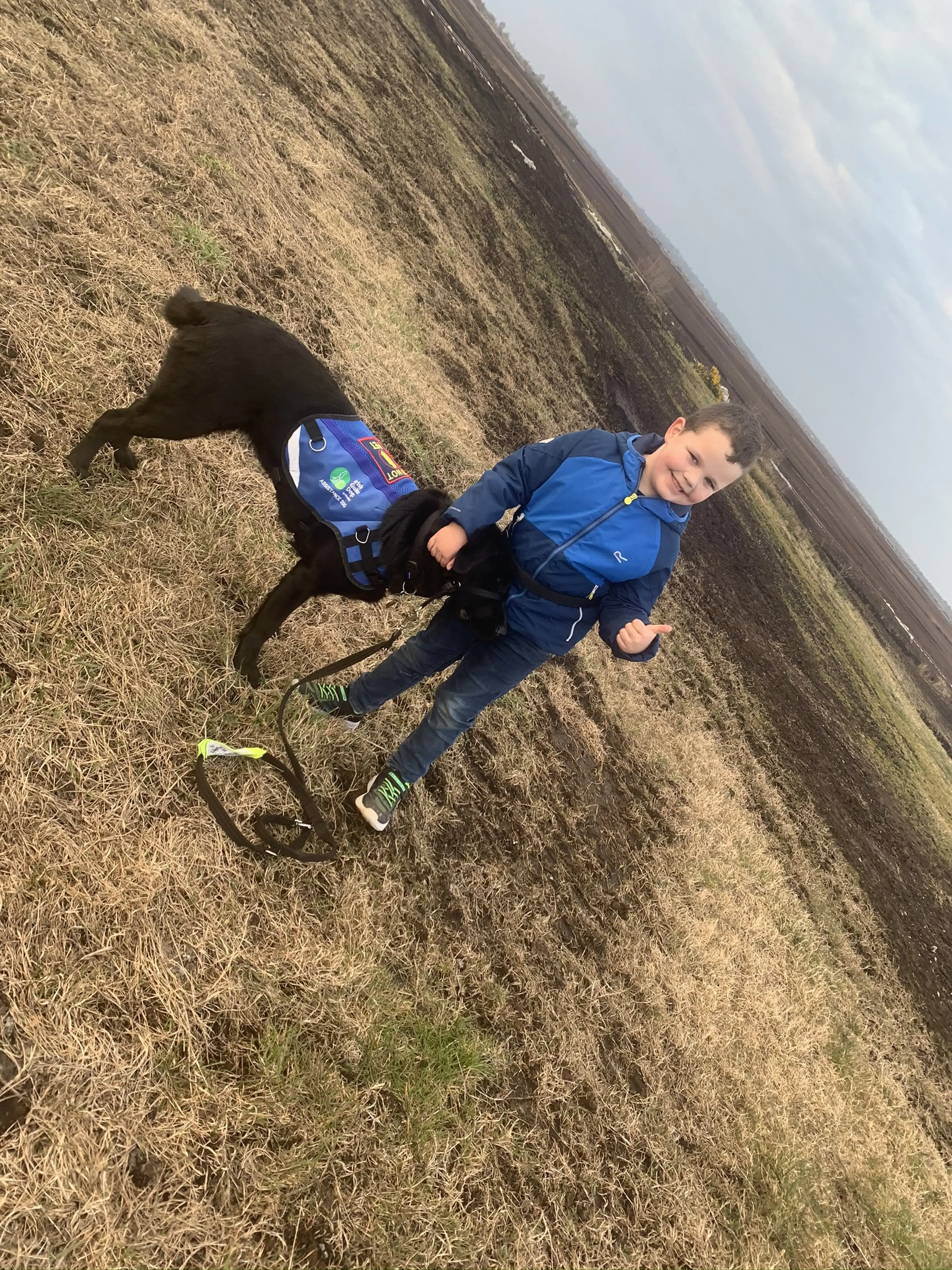 A young child in a blue jacket lies on a grassy hillside next to a black dog wearing a blue harness, both smiling at the camera.