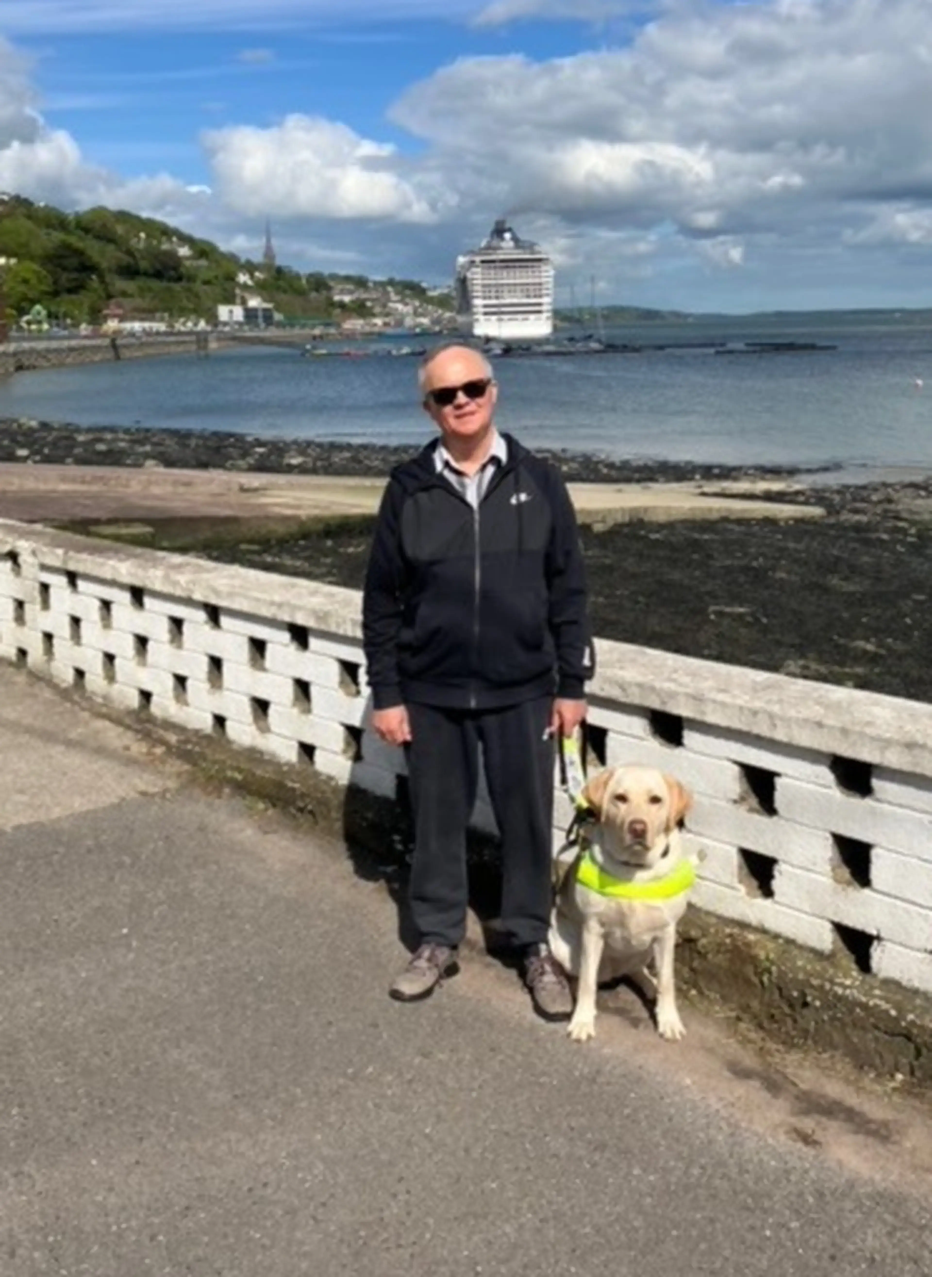 A man wearing sunglasses and dark athletic clothing stands with a light-colored Labrador retriever wearing a yellow safety vest on a coastal promenade, with a large cruise ship doc