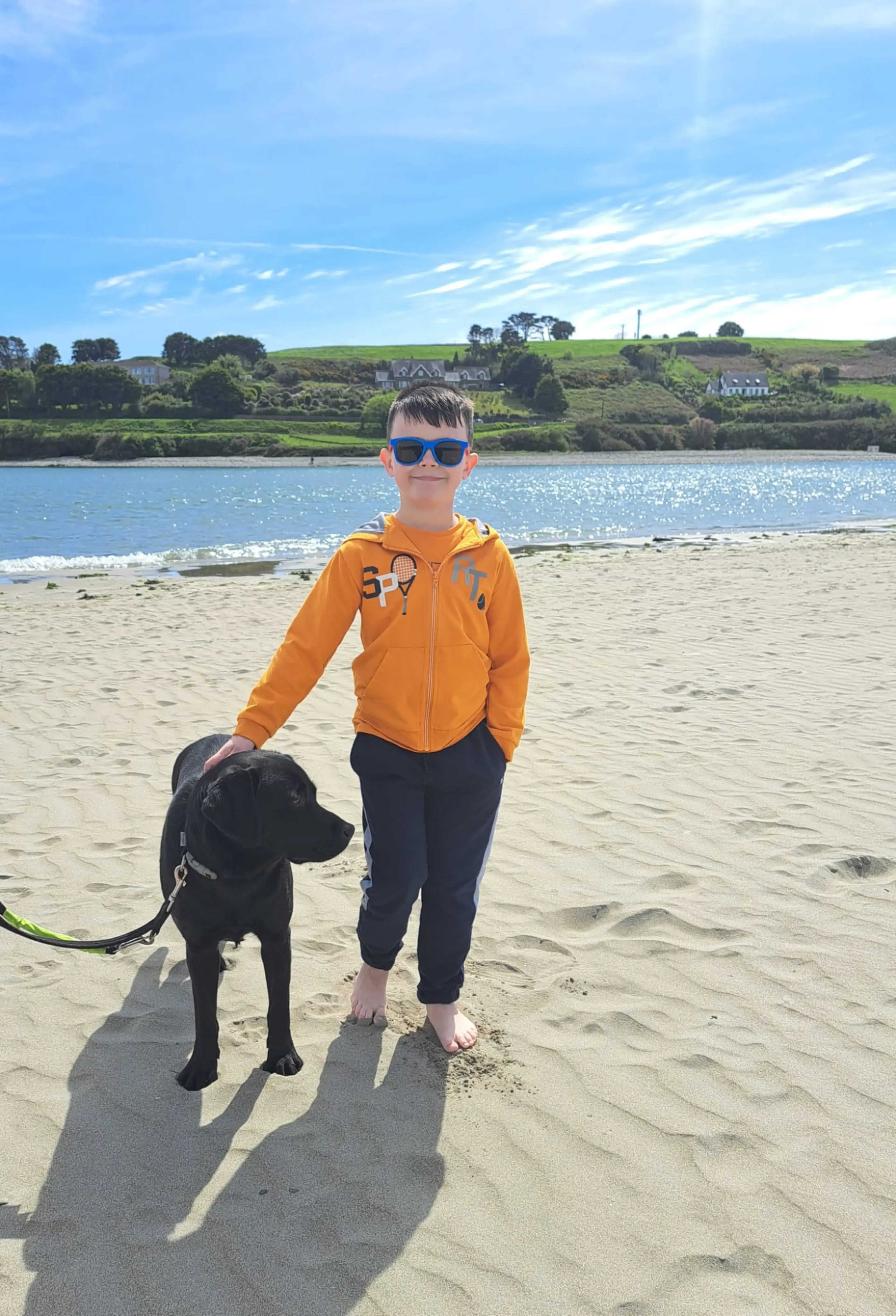 A young boy wearing sunglasses and an orange hoodie stands on a sandy beach holding the leash of a black dog, with rolling green hills and houses visible across the water behind hi