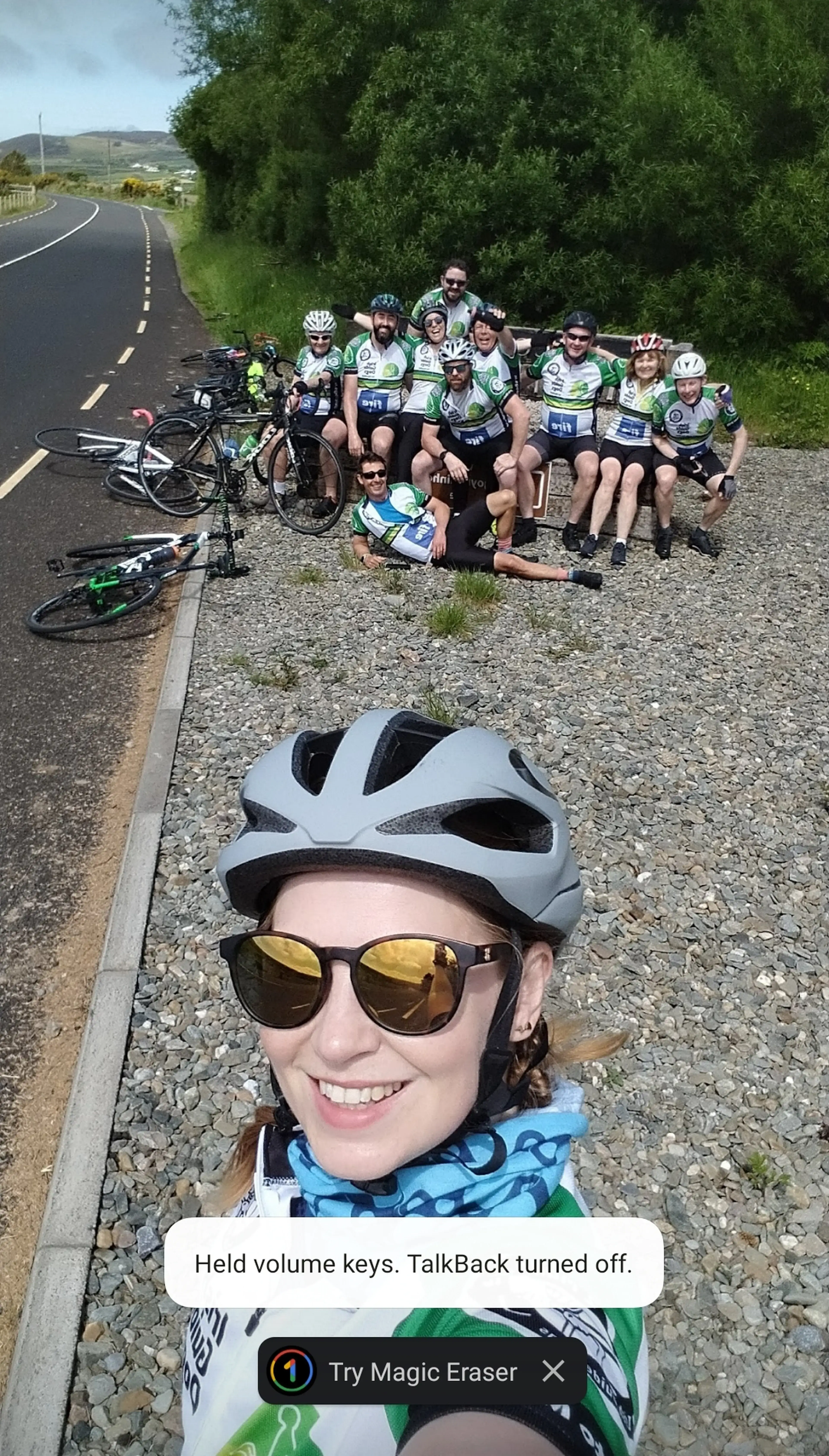 A cyclist in a white helmet and yellow-tinted sunglasses takes a selfie on a rural road while a group of cyclists in green and white team jerseys pose together in the background.