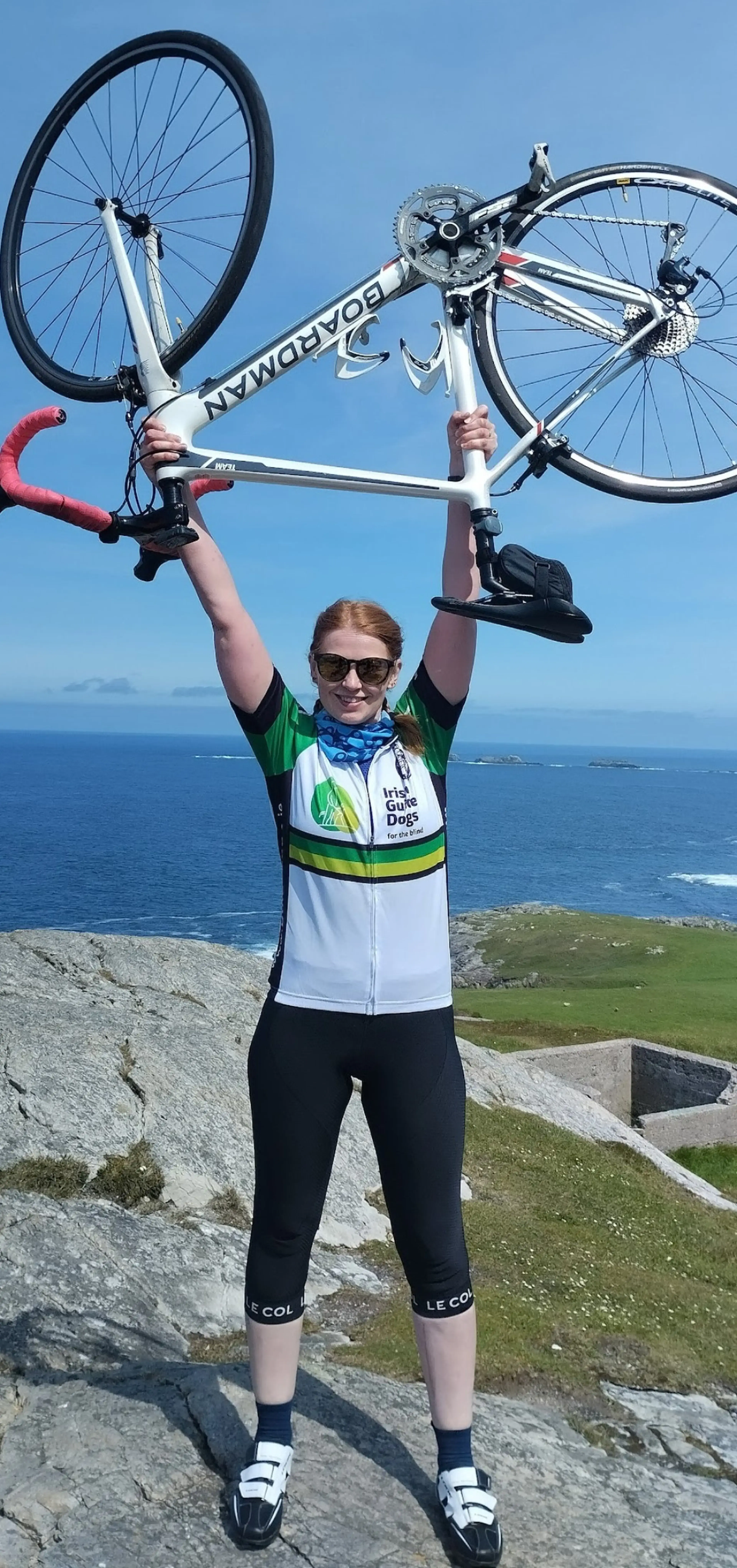 A cyclist in Irish Guide Dogs jersey holds her bike overhead while standing on rocky coastal terrain with the ocean and green landscape visible behind her.