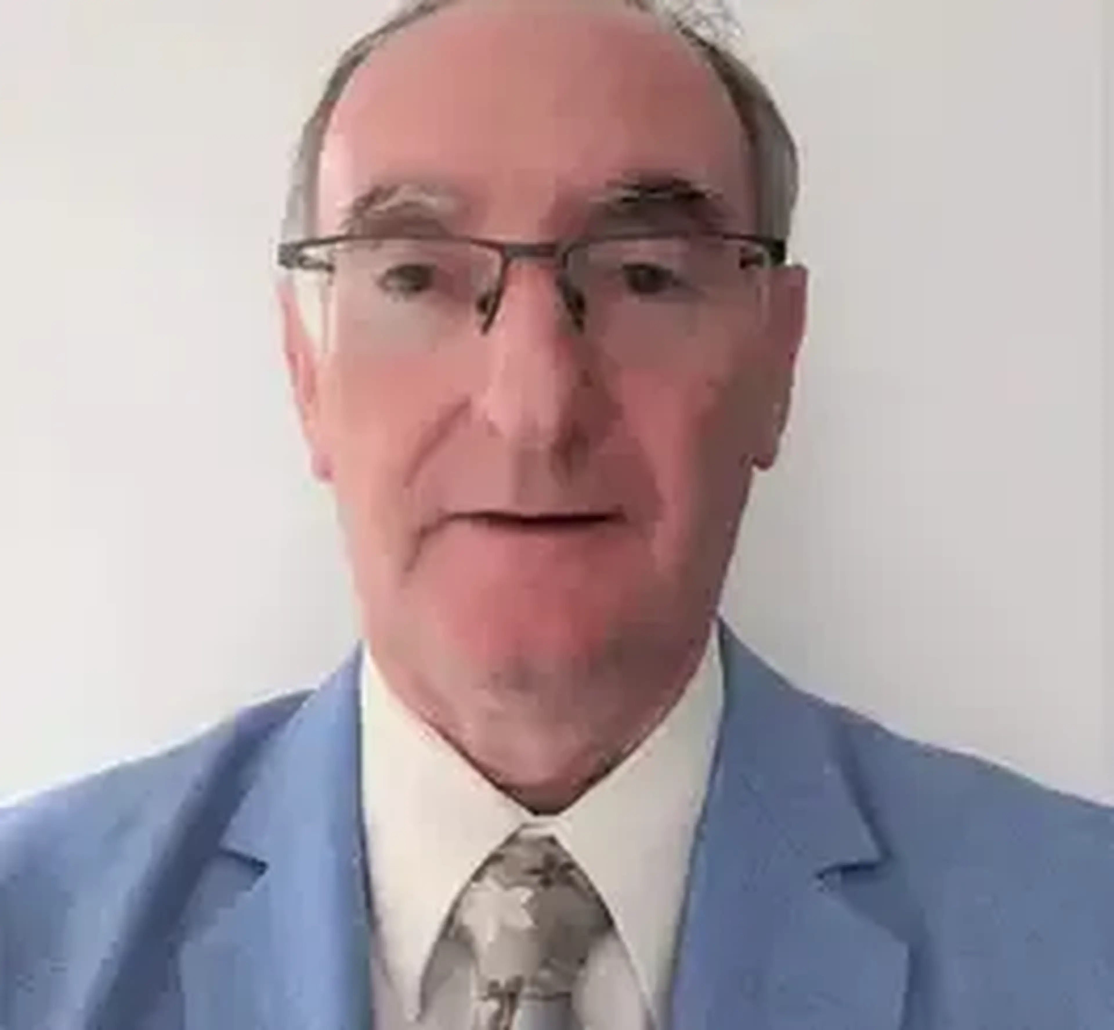A middle-aged man wearing glasses, a light blue blazer, and a patterned tie looks directly at the camera in a professional headshot.