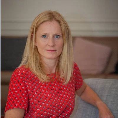 The image shows a middle-aged woman with long, blonde hair wearing a bright red patterned blouse and sitting on a couch.