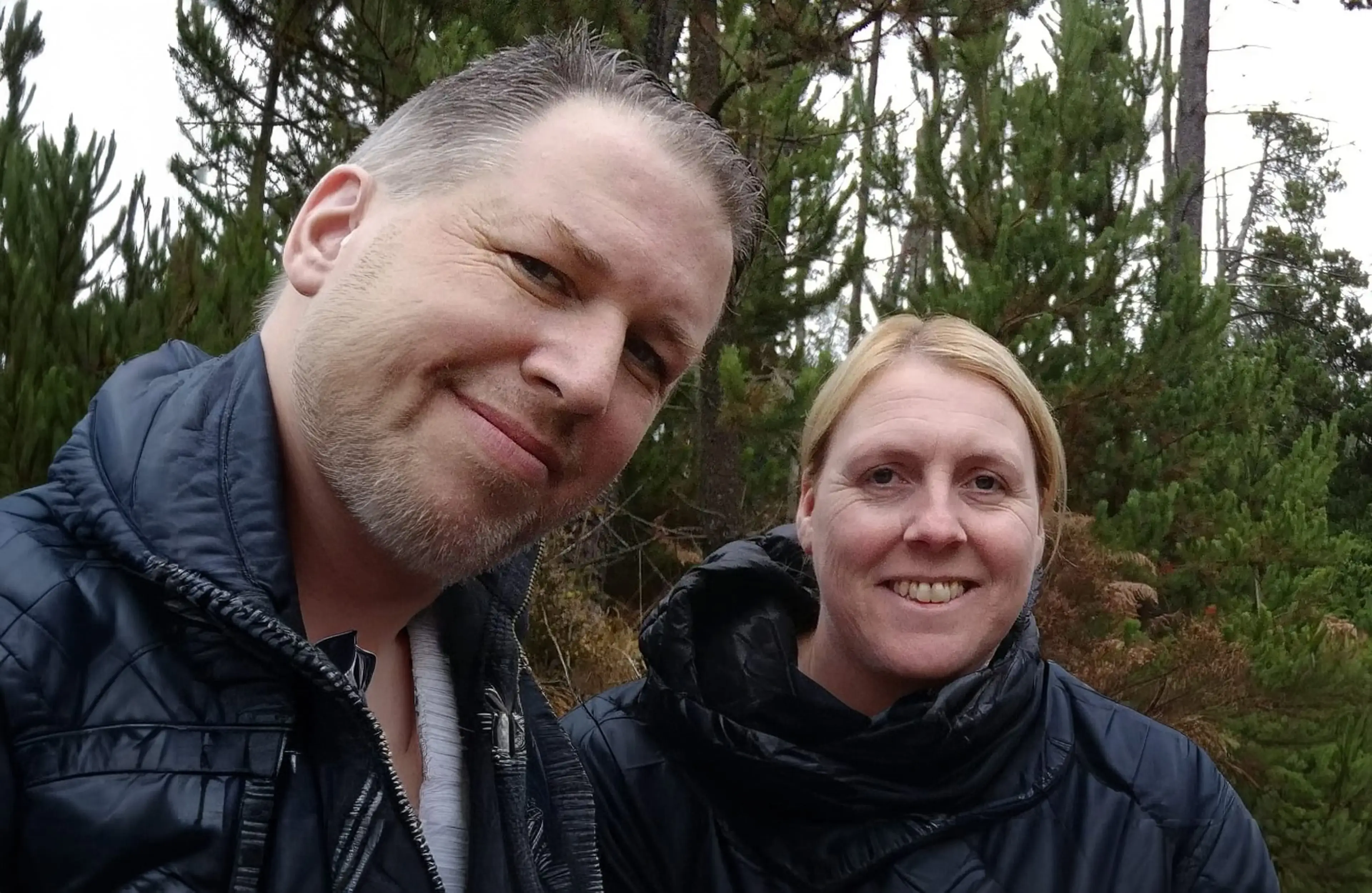 Two people smile at the camera while standing outdoors in front of tall evergreen trees on an overcast day.