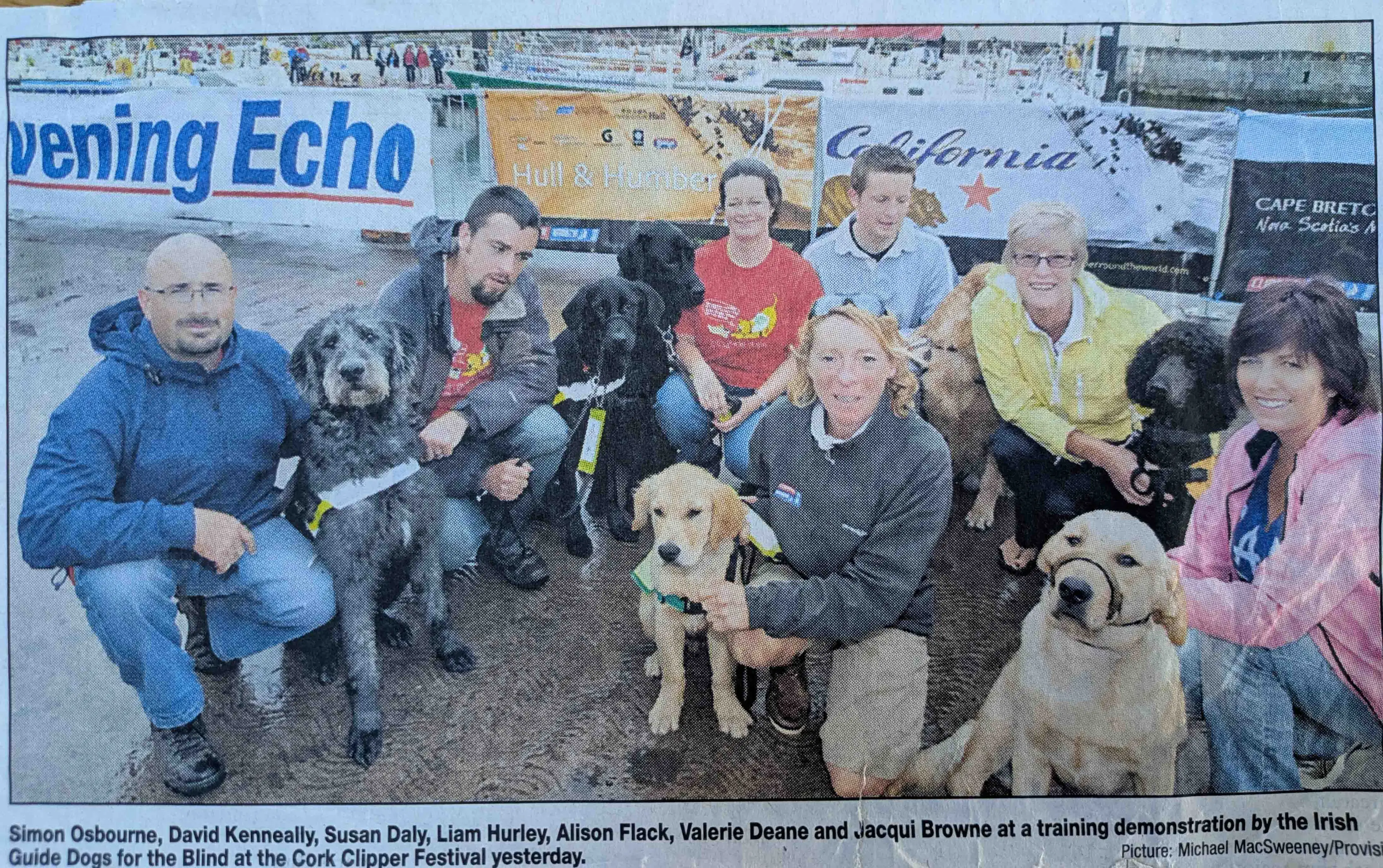 # Alt Text A group of people pose with six guide dogs of various colors at a training demonstration at the Cork Clipper Festival, with boats and harbor visible in the background.
