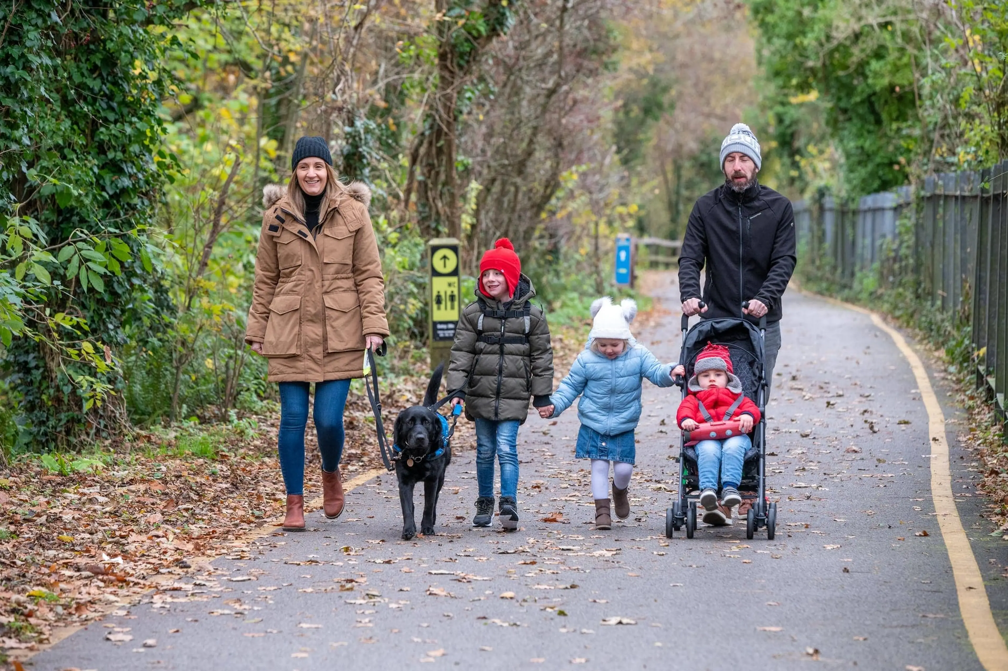 A family of five, including two adults and three children, walks along a tree-lined path with a black dog on a leash during autumn, all wearing warm clothing.