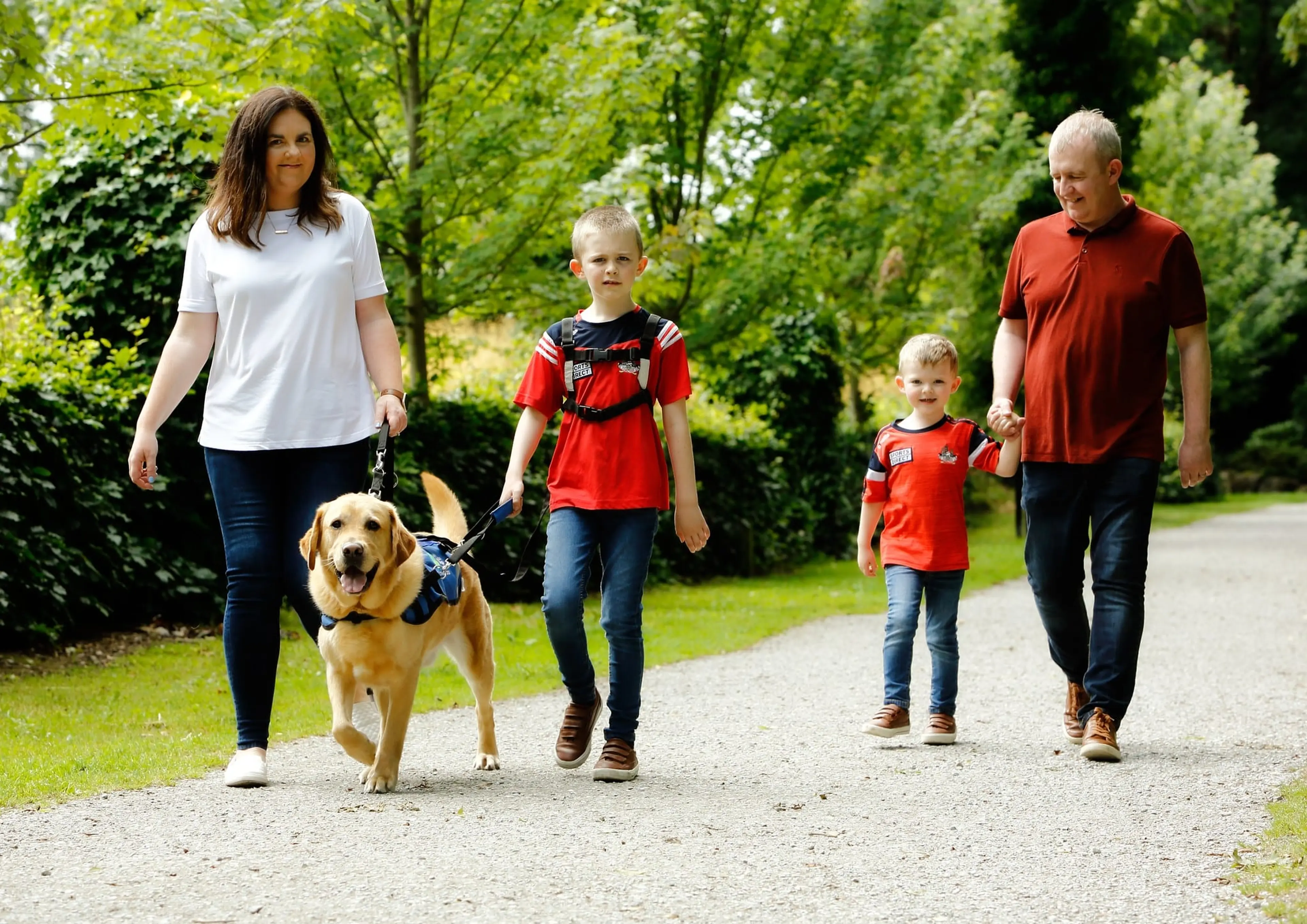 A family of four, including a woman, two young boys, and a golden retriever, are walking together on a gravel path through a lush, green park.