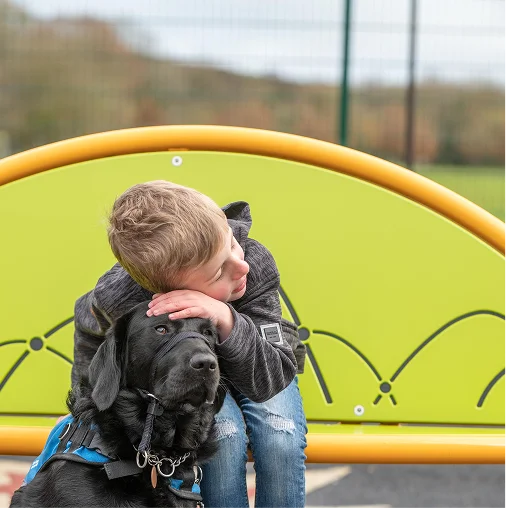 A young child hugs a black dog from behind while sitting in front of a bright yellow-green playground structure.