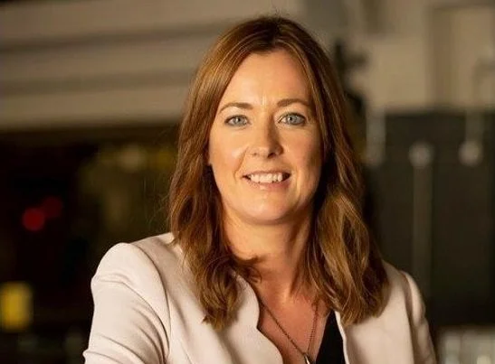 The image shows a smiling woman with long brown hair wearing a beige top.