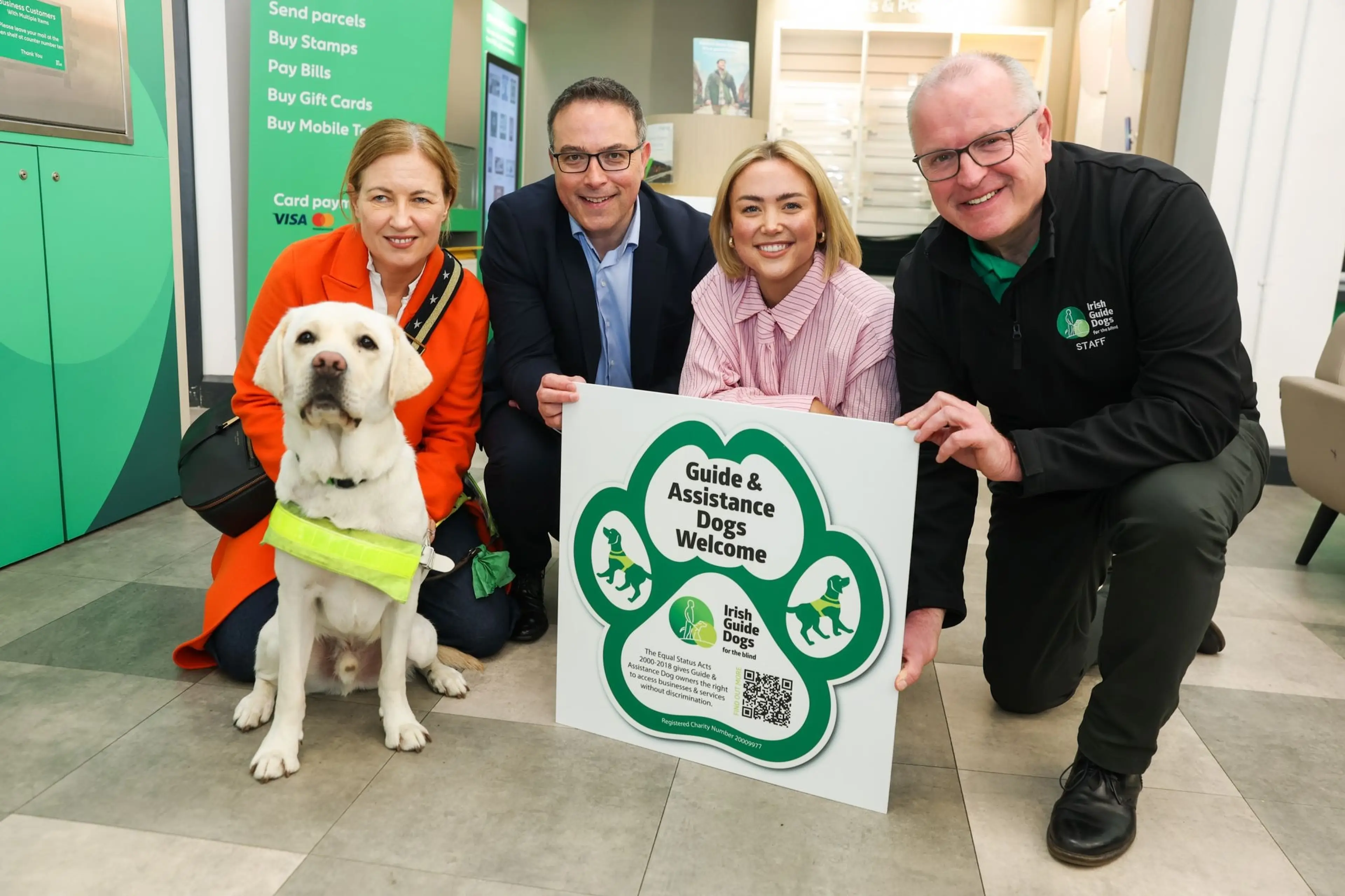 The image shows a group of people, including a guide dog and staff members from the Irish Guide Dogs, standing together and holding a sign that welcomes guide and assistance dogs.