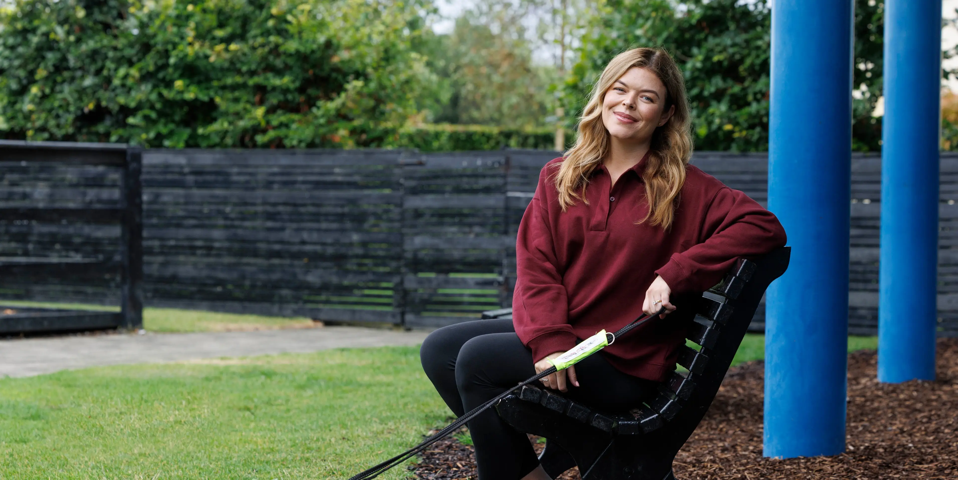 A smiling woman sits on a park bench holding a walking stick, with trees and a fence in the background.