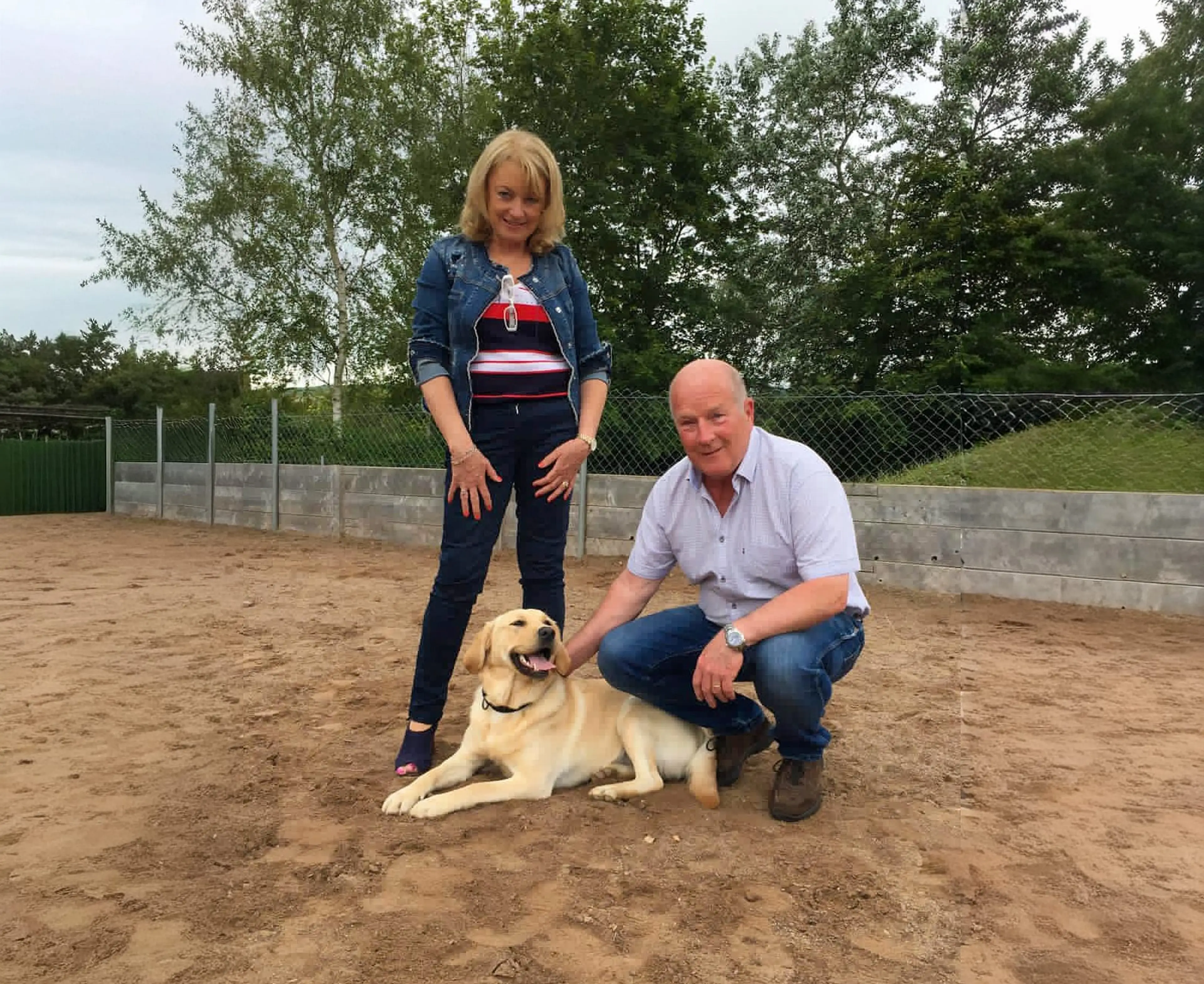 A man and woman pose with a yellow Labrador Retriever in a sandy arena surrounded by fencing and trees.