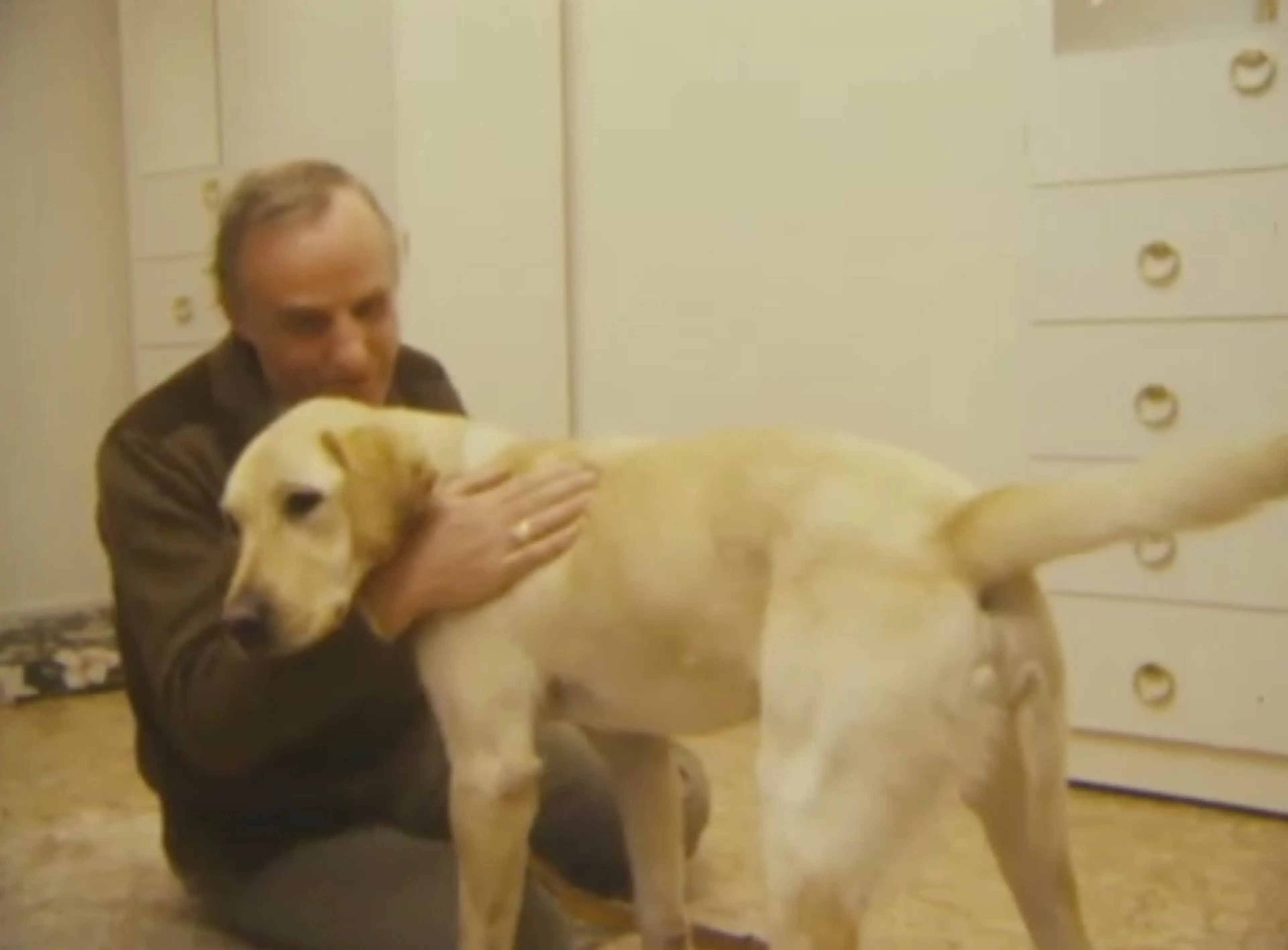 An elderly man wearing dark clothing is embracing a large, light-colored dog in an indoor setting with a plain background.