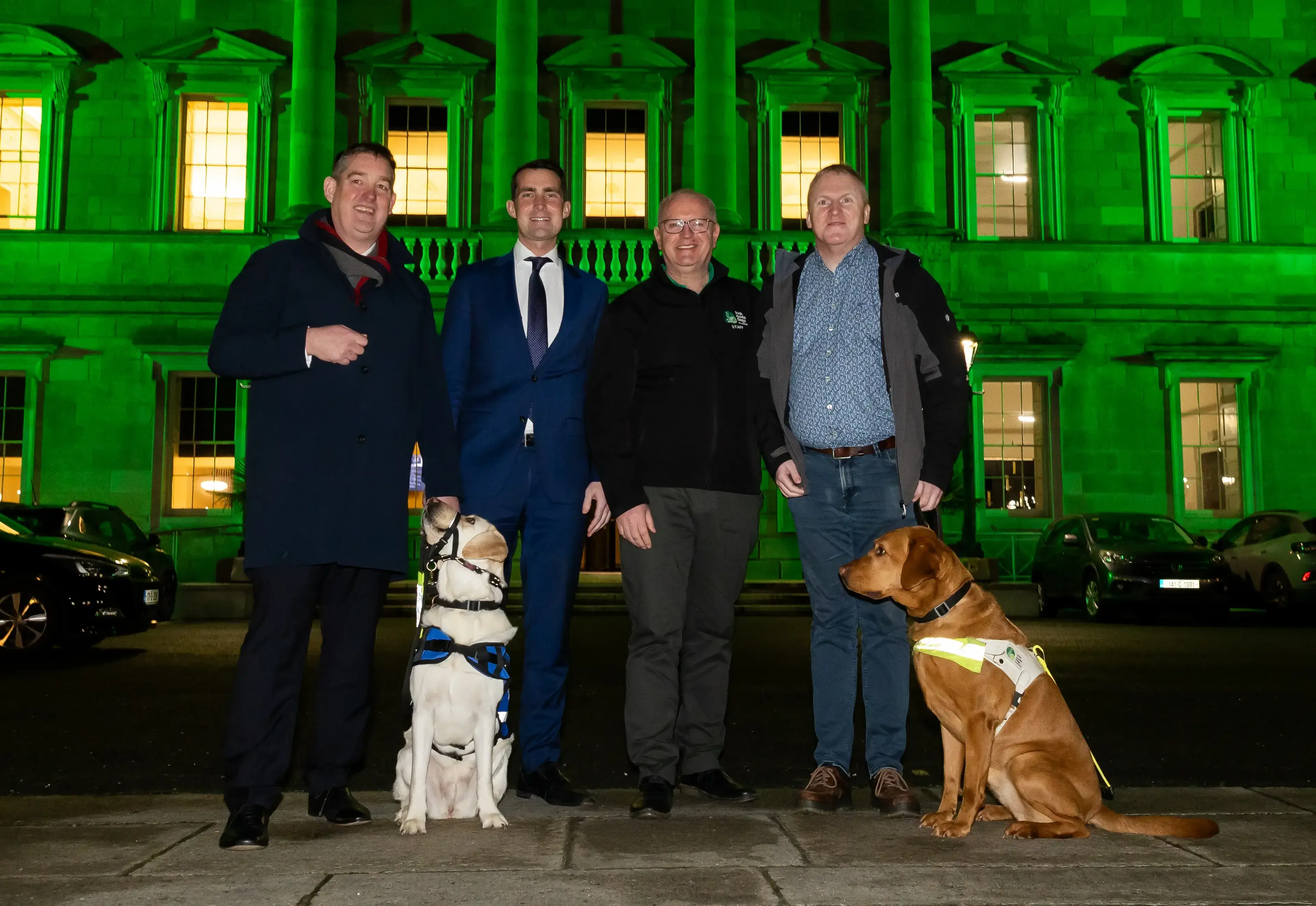 Four men stand with two service dogs in front of an illuminated green-lit historic building at night.