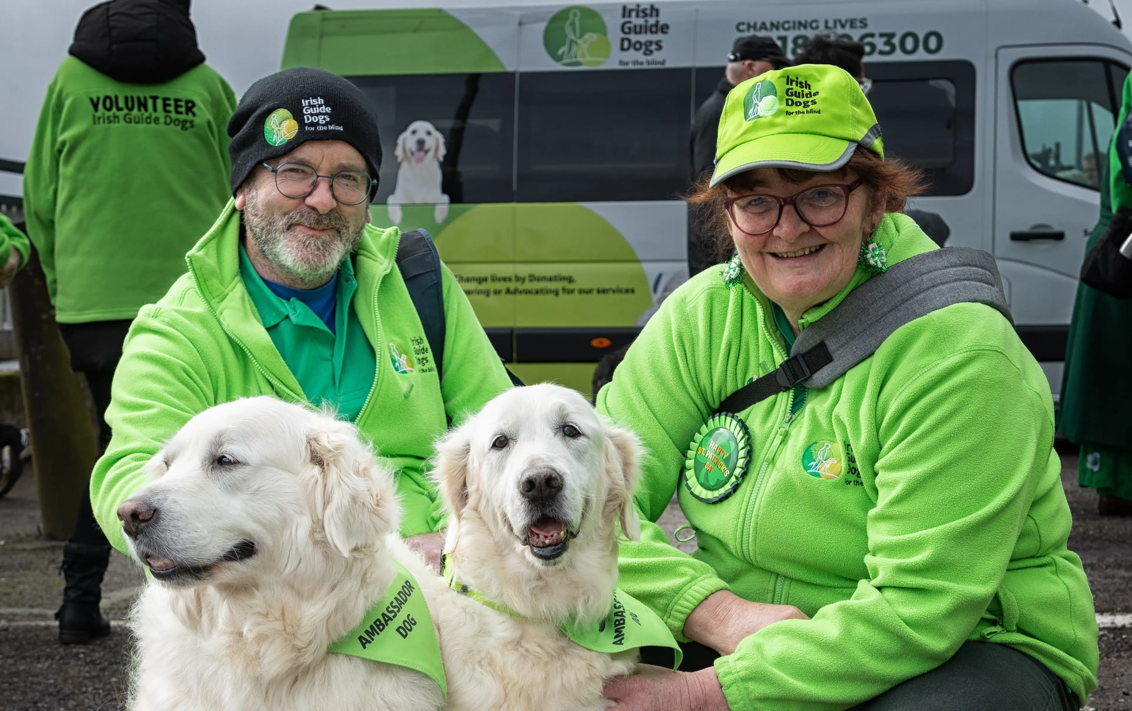 The image shows two volunteers from the Irish Guide Dogs organization, wearing bright green vests and hats, posing with two smiling service dogs wearing matching green vests.