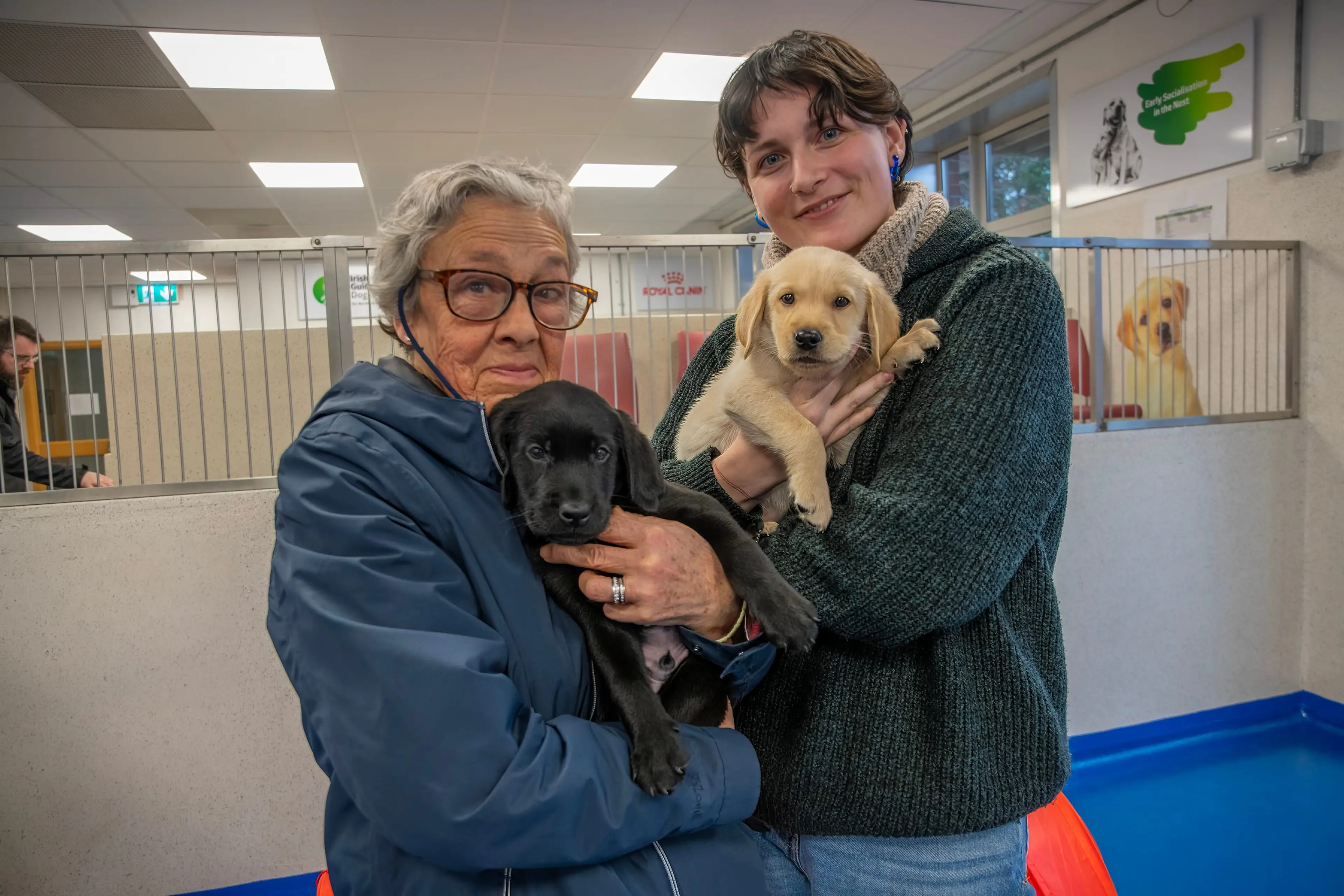 Two women, one older and one younger, are holding two puppies in an animal shelter or adoption center.