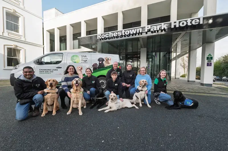 A group of people and multiple trained service dogs pose together in front of the Rochestown Park Hotel, with a van displaying "Irish Guide Dogs" and "Changing Lives" messaging vis