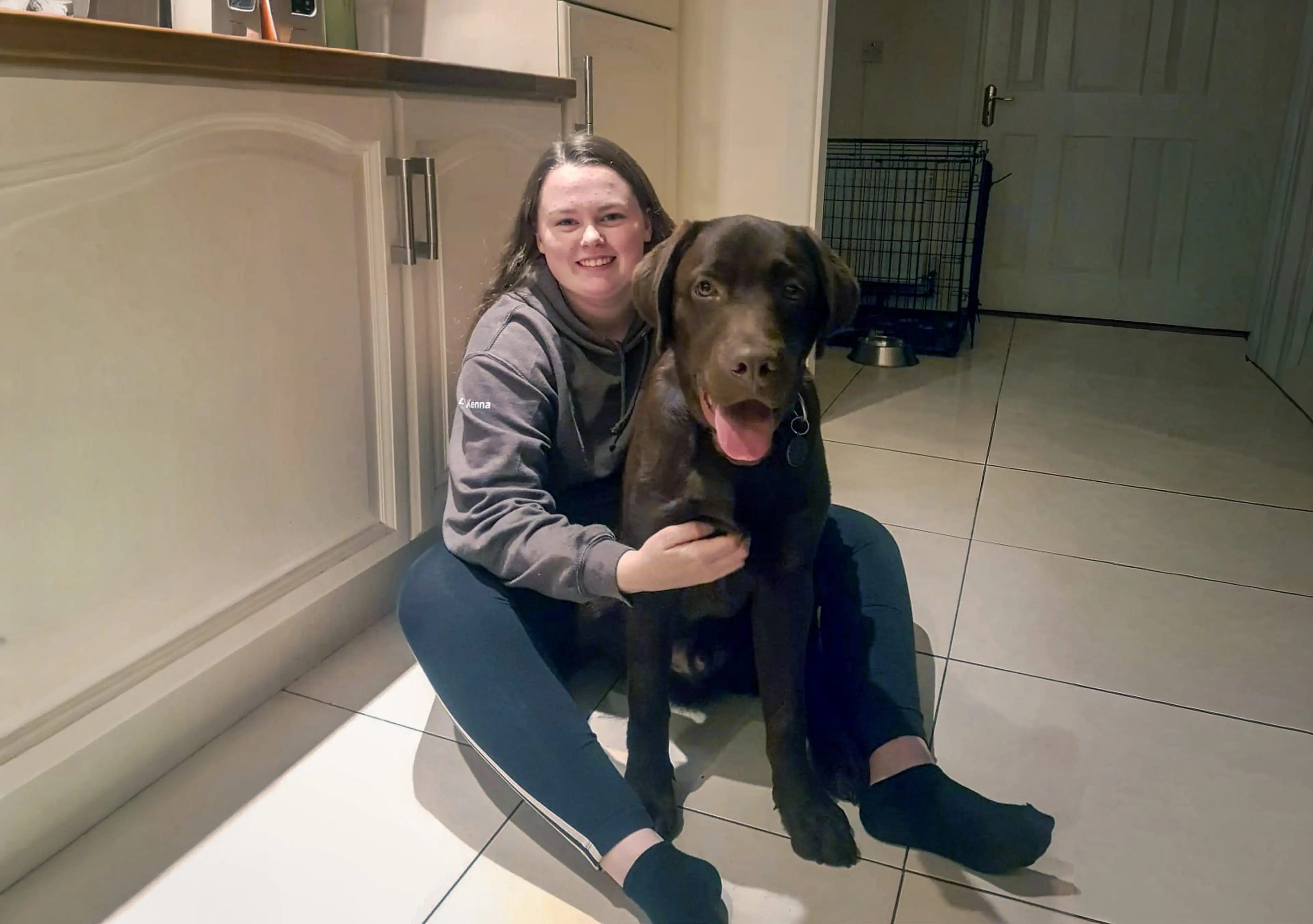 A smiling young woman crouches beside a happy brown Labrador Retriever in a tiled kitchen or utility room.