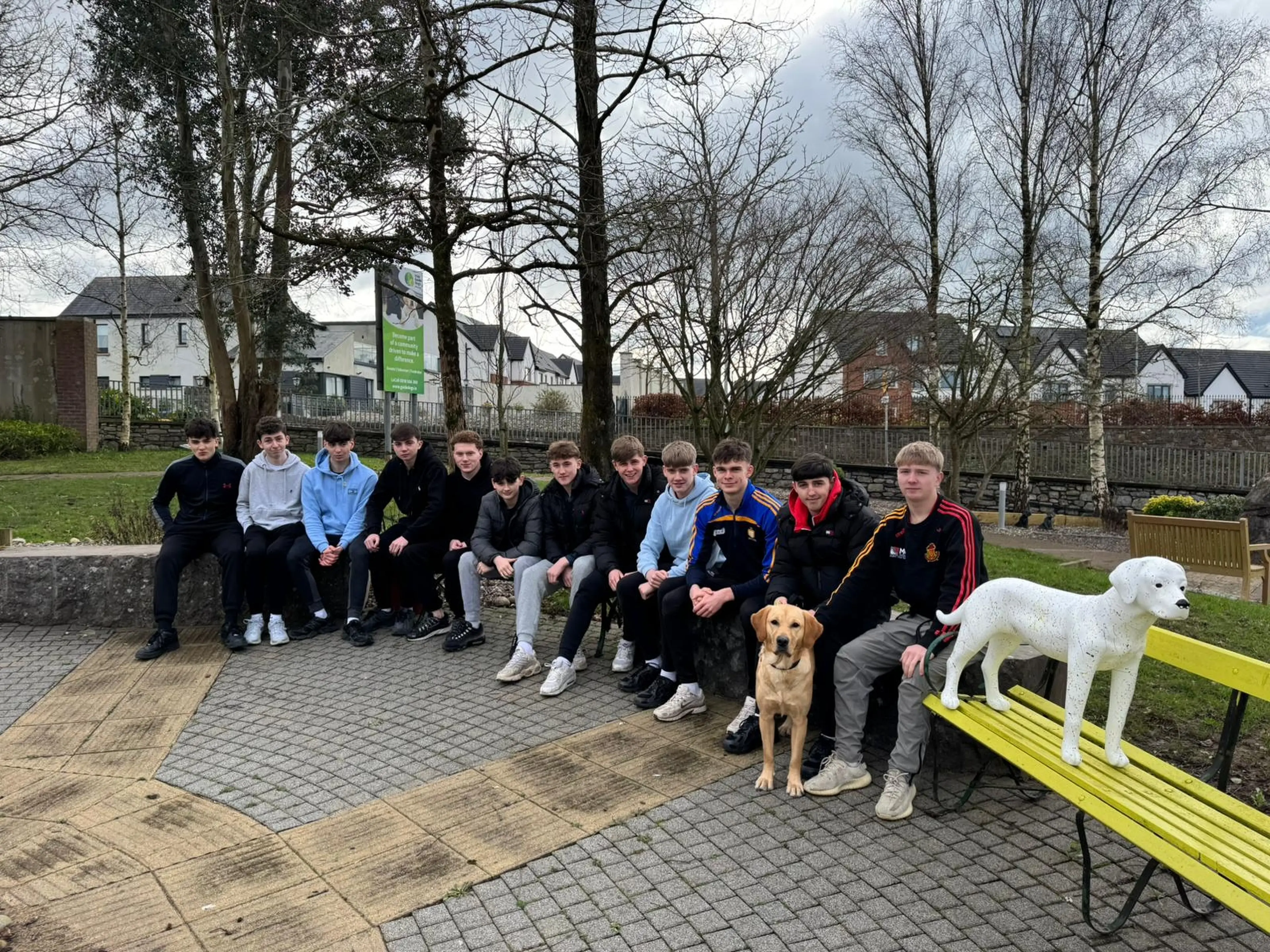 The image shows a group of young people, both boys and girls, sitting together on a bench in a park-like setting, with a dog and a white statue-like figure nearby.